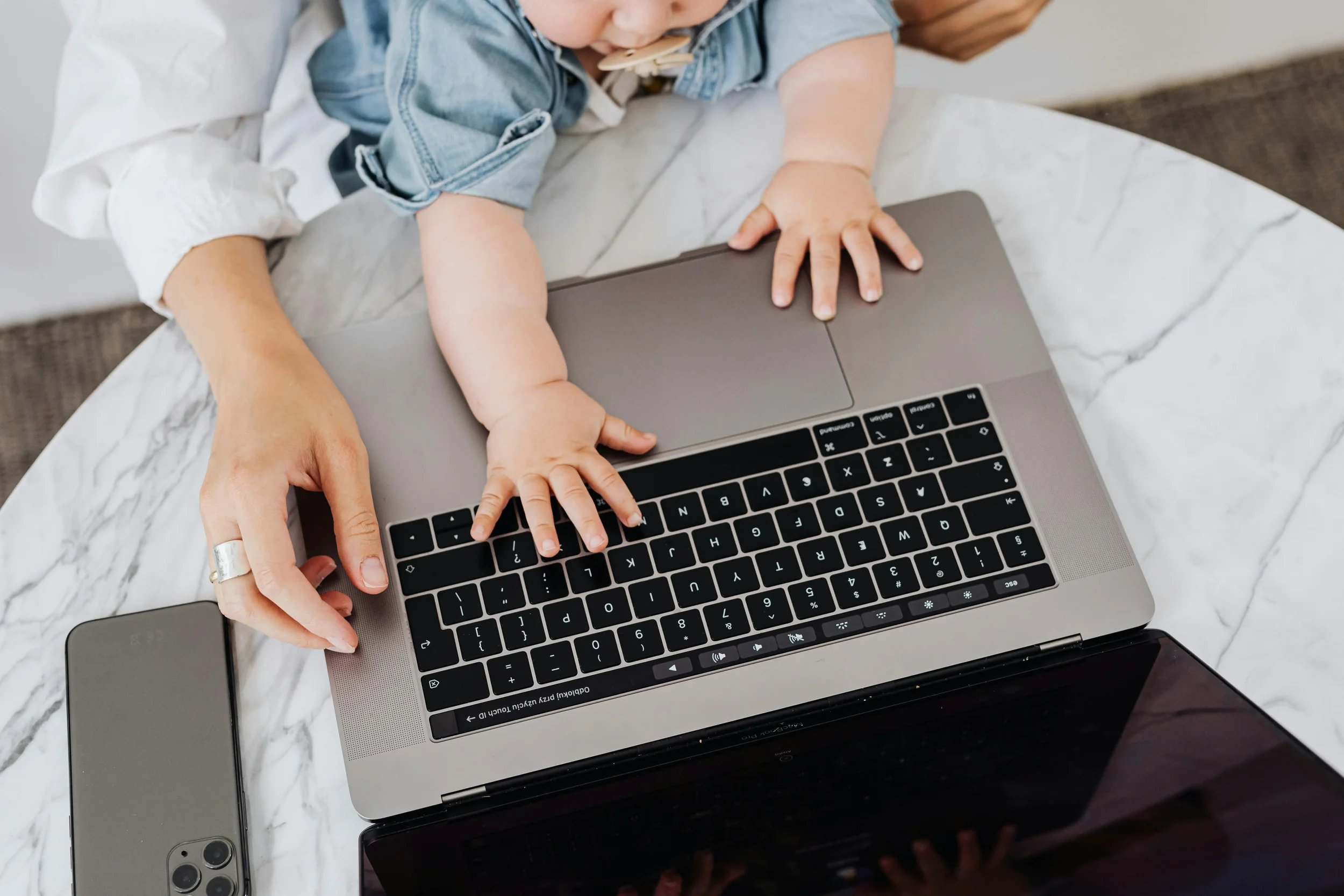Person assisting a child with typing on a laptop keyboard, with a smartphone nearby on a marble table.