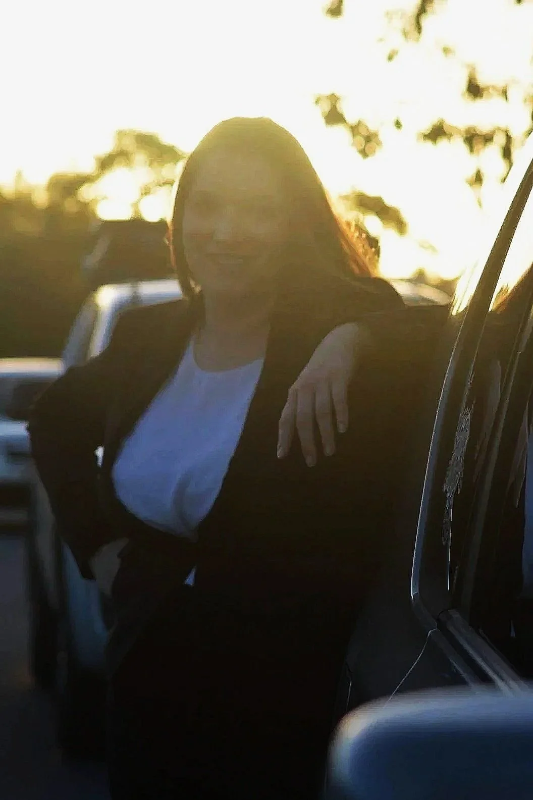 A woman leaning against a car with her arm resting on it, outside during sunset, with trees in the background.