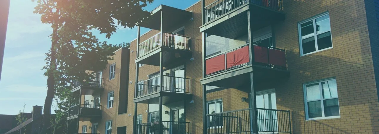 Multistory apartment building with multiple balconies, some with outdoor furniture and red privacy screens, brick exterior, and large windows, with a large tree in front.