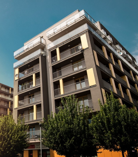 Multi-story apartment building with balconies, surrounded by green trees, under a clear blue sky.