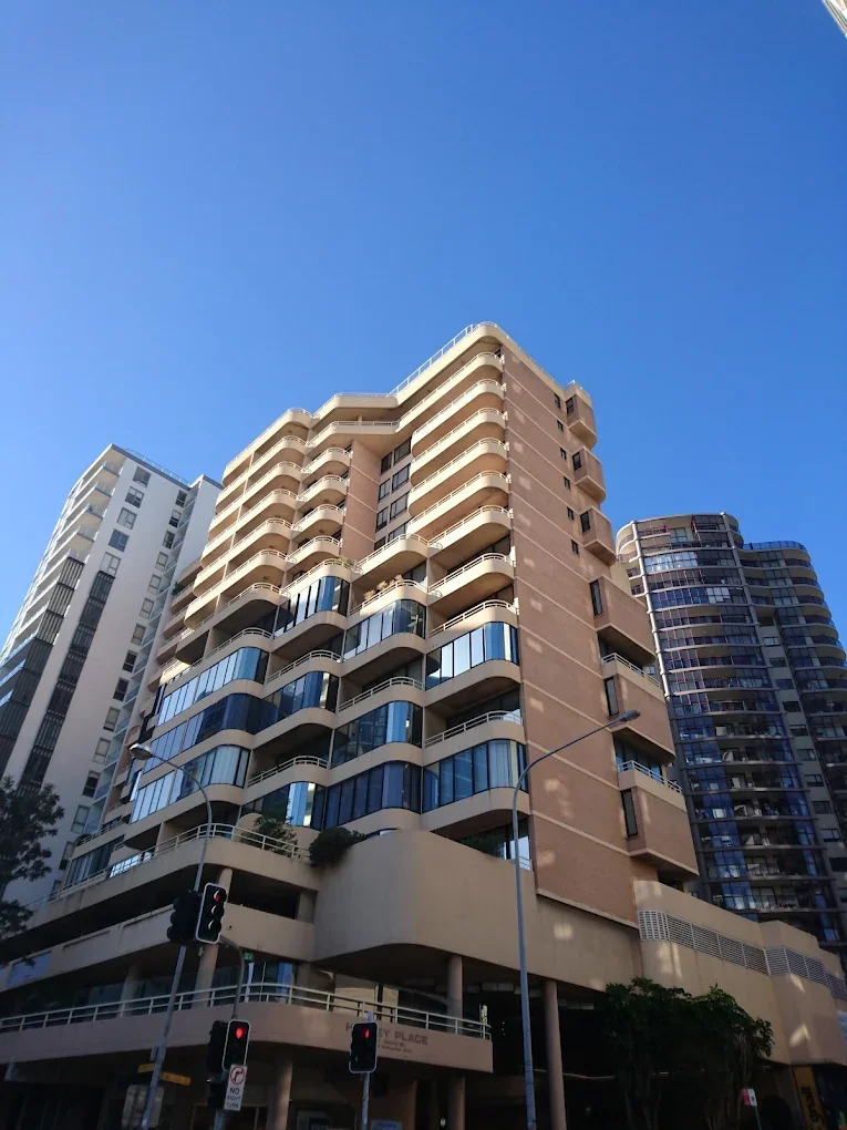 A tall modern apartment building with curved balconies and glass windows, situated in an urban area on a sunny day with clear blue sky.