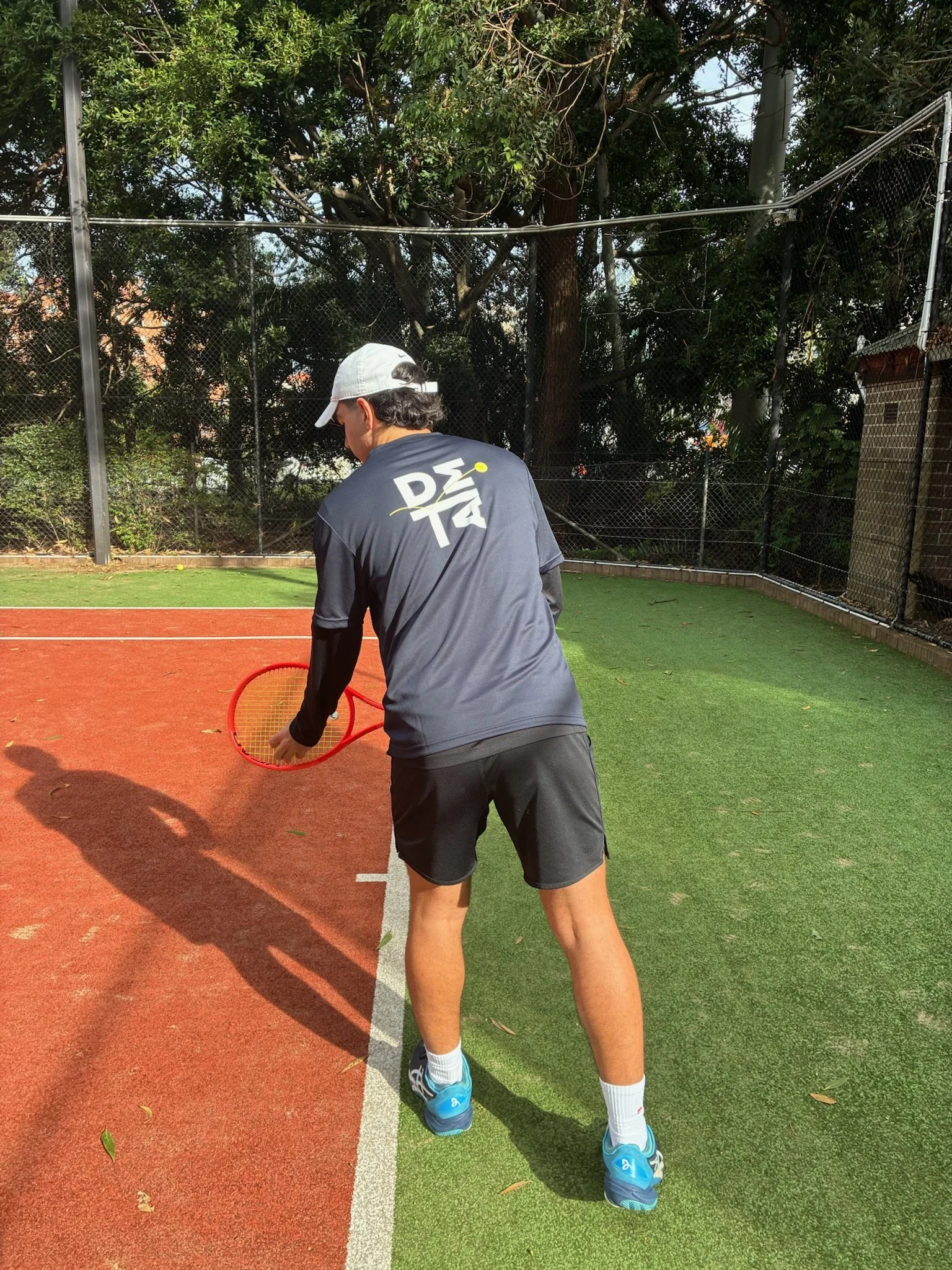 A person on a tennis court holding a red tennis racket, wearing a gray Nike cap, gray Nike t-shirt, black shorts, white socks, and blue sports shoes, with the court marked by white lines.