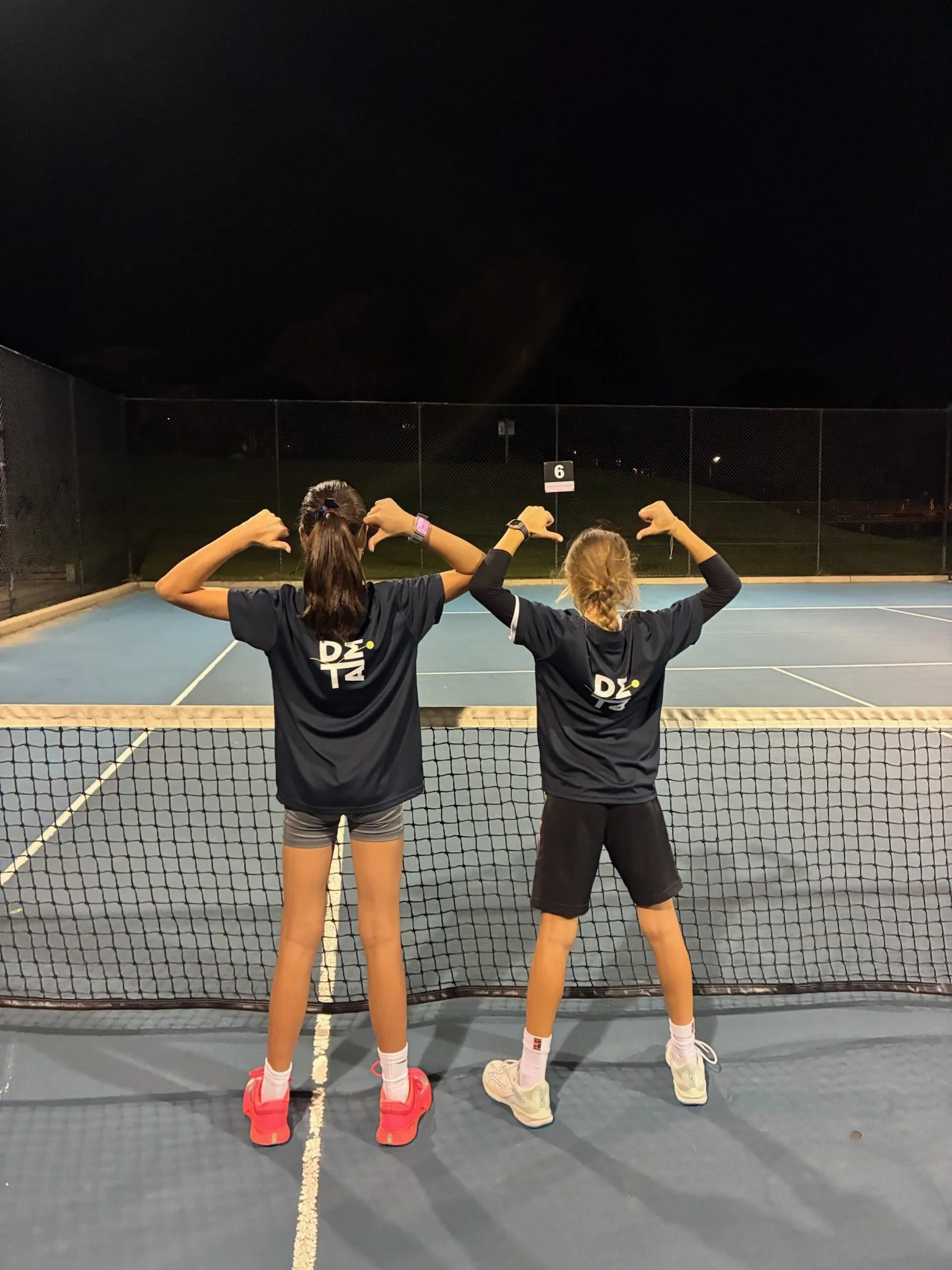 Two young girls stand side by side on a tennis court at night with their backs to the camera, flexing their muscles and showing strength. The court is enclosed with a chain-link fence and is lit by artificial lighting, with a dark sky in the background.