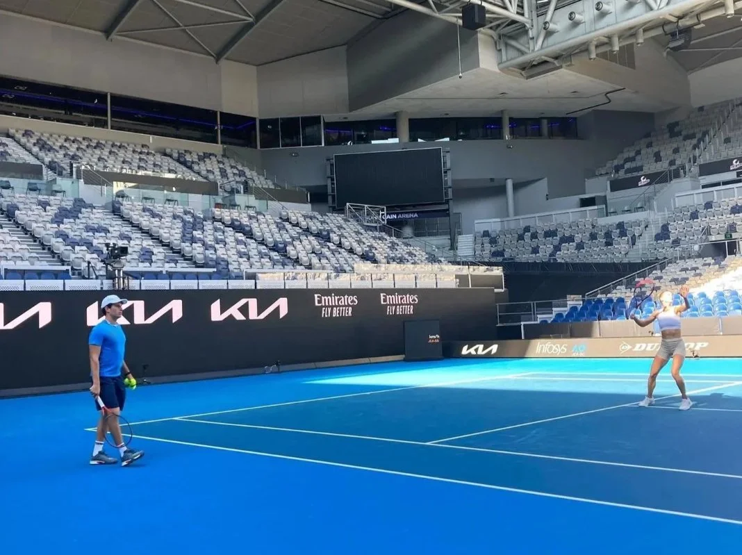 Two people on a blue tennis court practicing serving, with empty stadium seats and advertising banners in the background.