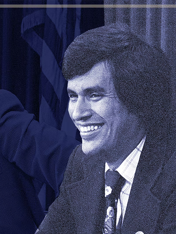 A man smiling and waving, dressed in a suit and tie, with an American flag in the background.
