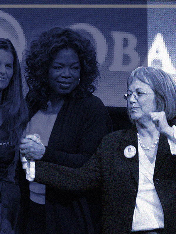 Three women on stage during a political event, with two women clasping hands and the third woman looking on.