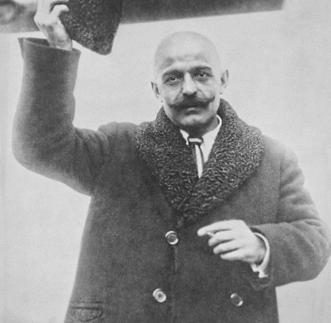 A man with a bald head and a mustache, wearing a coat with a fur collar, holding a hat in his right hand, and pointing with his left hand.