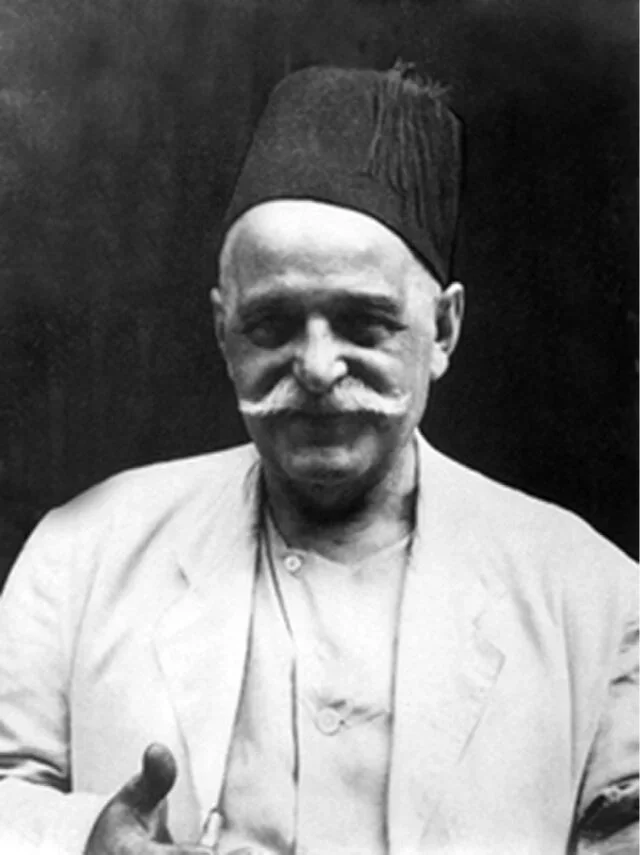 A black-and-white photo of an older man with a mustache, wearing a chef's coat and a tall chef's hat, smiling and giving a thumbs-up.