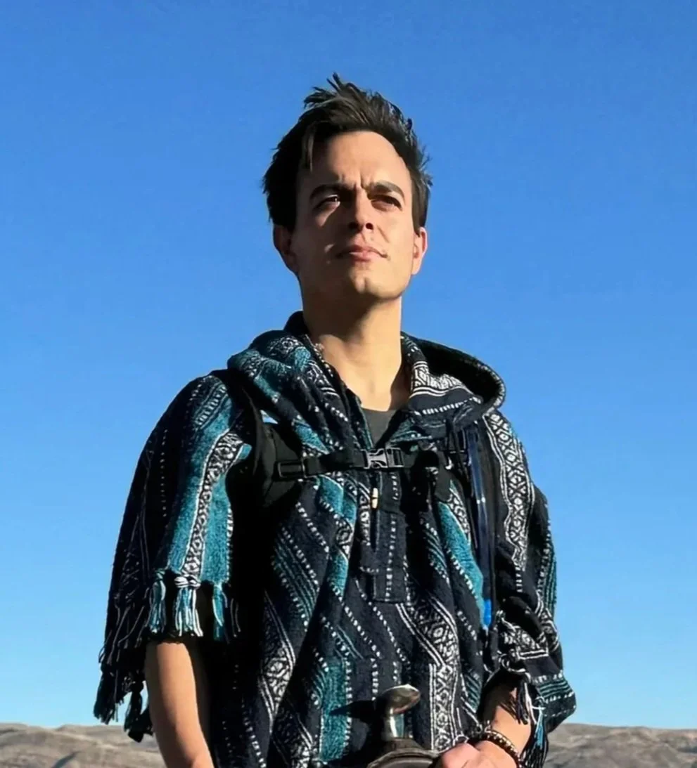A young man standing outdoors on a clear day with a blue sky in the background. He has short, dark hair and is wearing a patterned hoodie and a backpack.