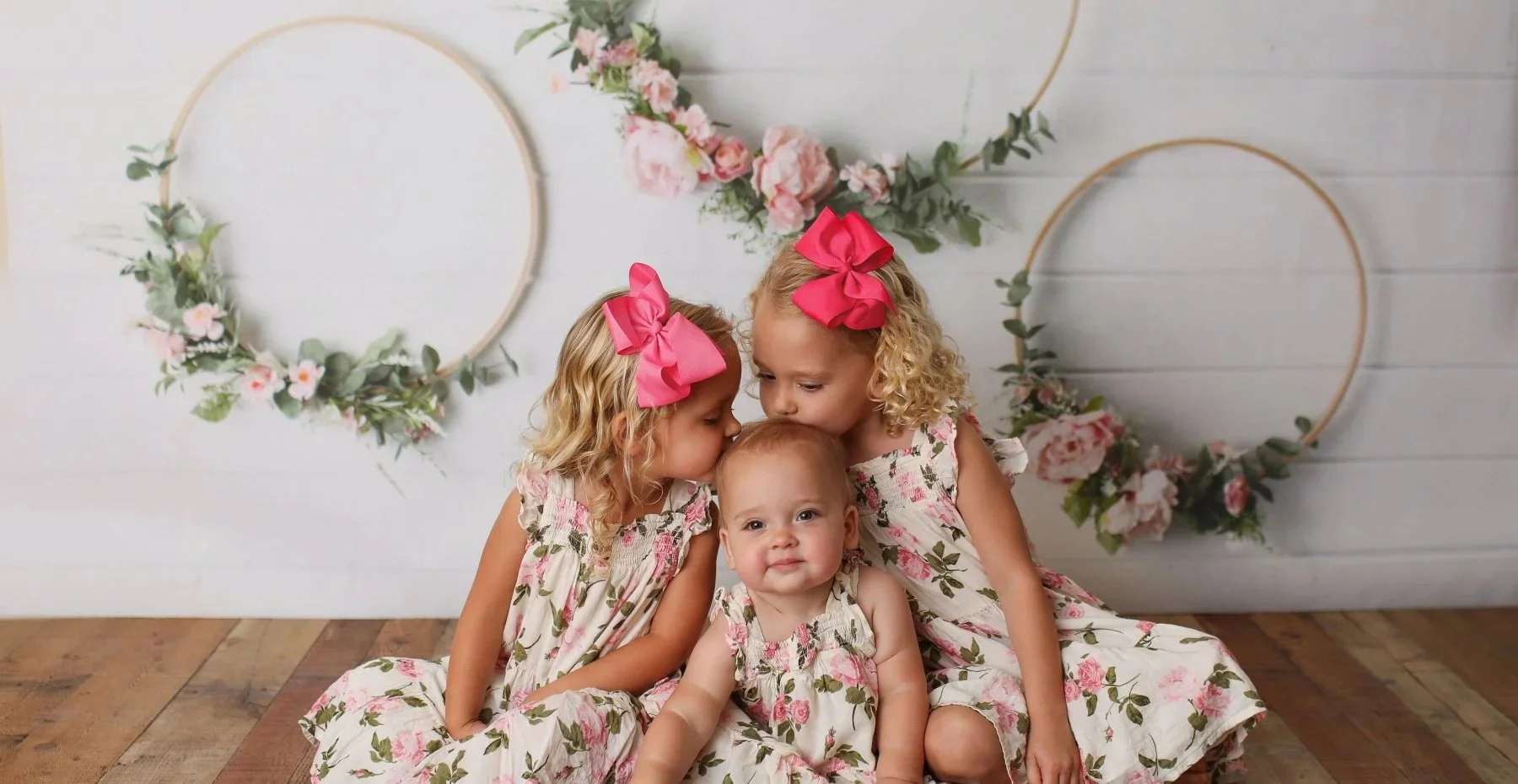Three young girls wearing floral dresses and pink bows sitting on a wooden floor in front of a white wall decorated with two floral wreaths.