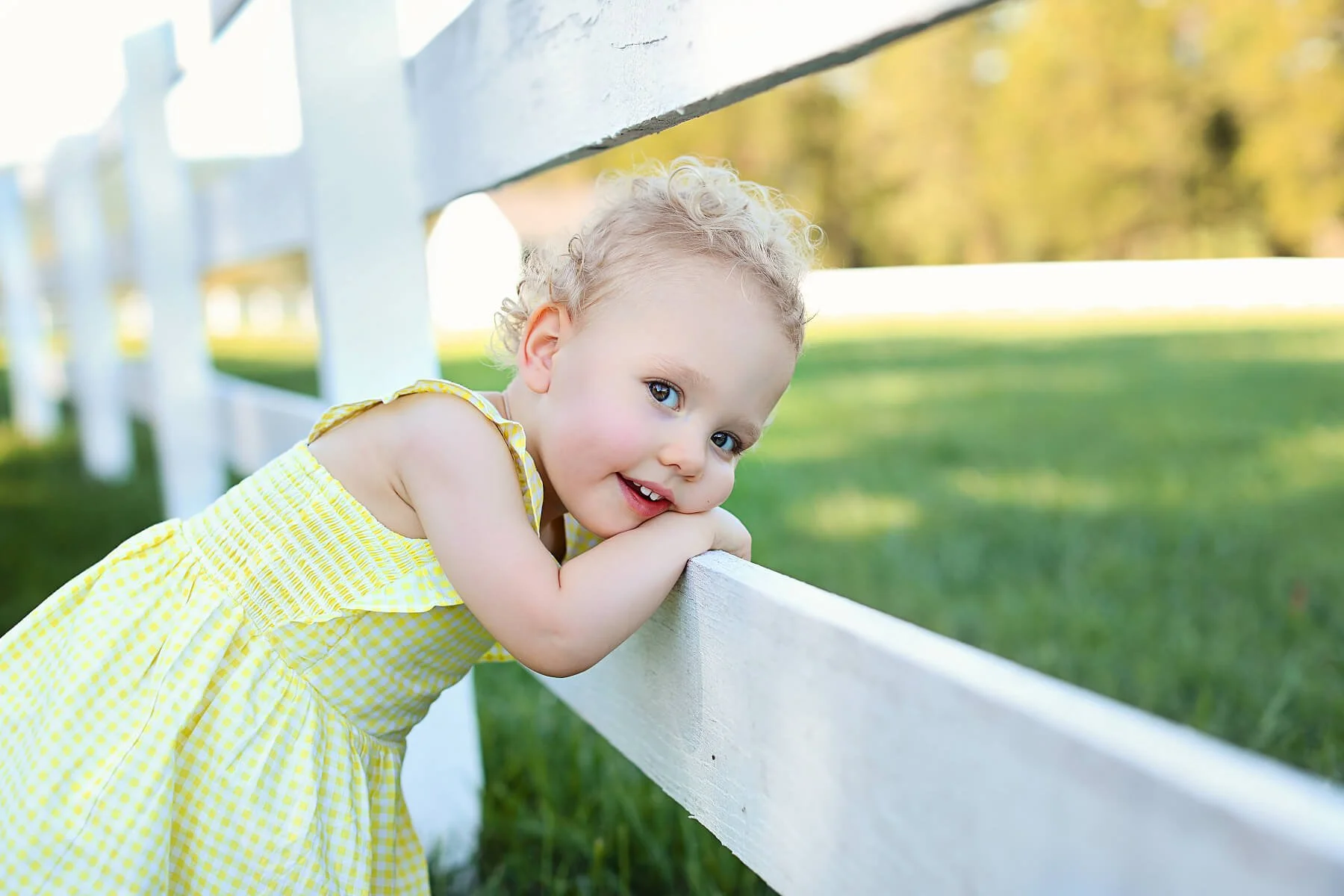 A young girl with curly blonde hair wearing a yellow checkered dress leaning on a white wooden fence outdoors, smiling and looking at the camera with a background of green grass and trees.