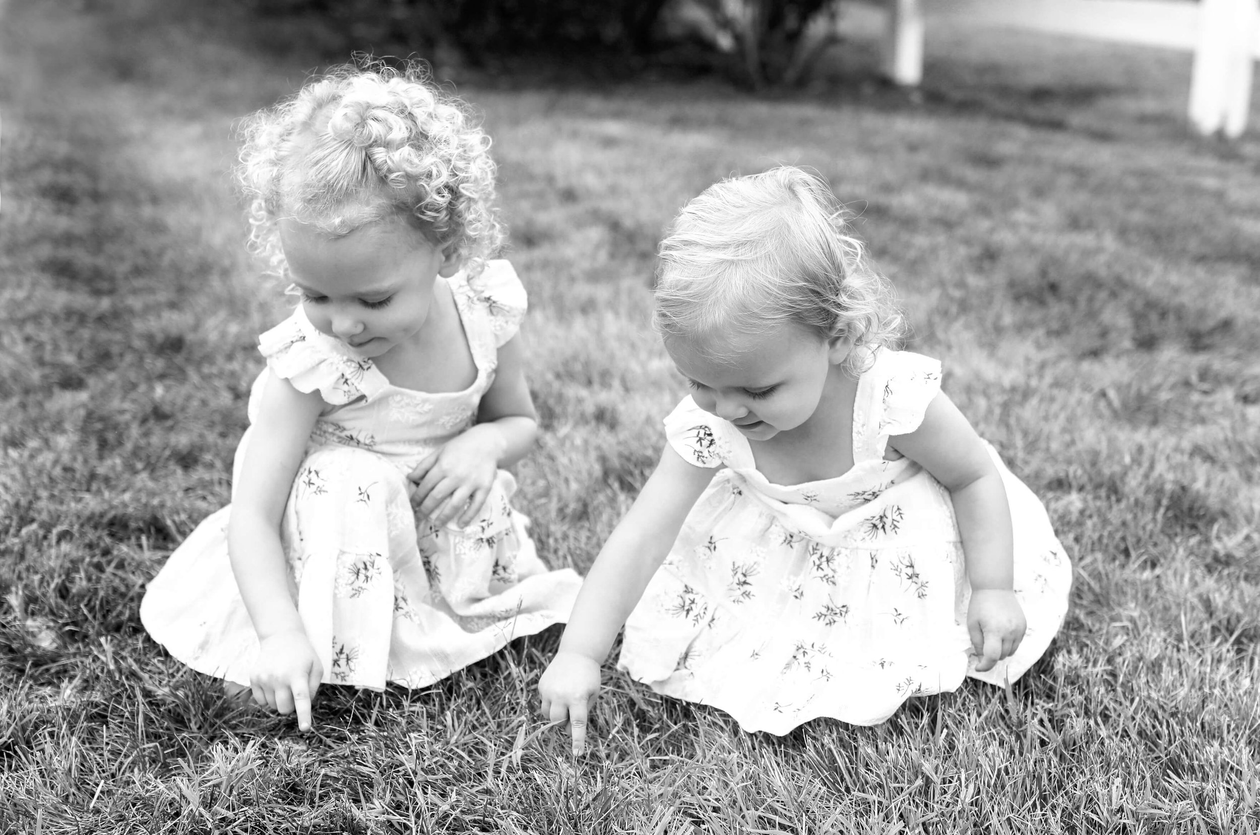 Two young girls with curly hair wearing white dresses picking grass in a grassy outdoor area.
