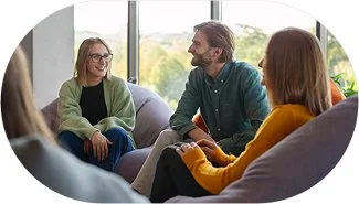 Group of four people sitting and talking in a bright room with large windows.