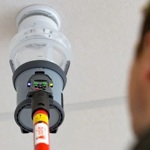 Close-up of a smoke detector with a test aerosol can directed at it, attached to a ceiling with part of a person's head visible in the foreground.
