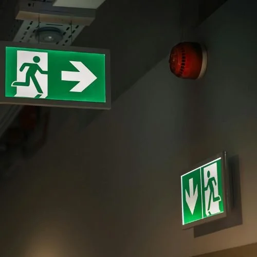 Green emergency exit signs, one with an arrow pointing right and a walking figure, and another with a downward arrow and a walking figure, on a gray ceiling with a red alarm.