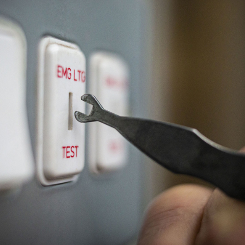 A person's hand holding a small tool pressing a switch labeled 'TEST' on a control panel.