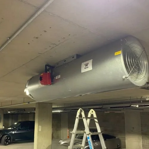 Large cylindrical industrial fan or heater mounted on the ceiling of a parking garage.