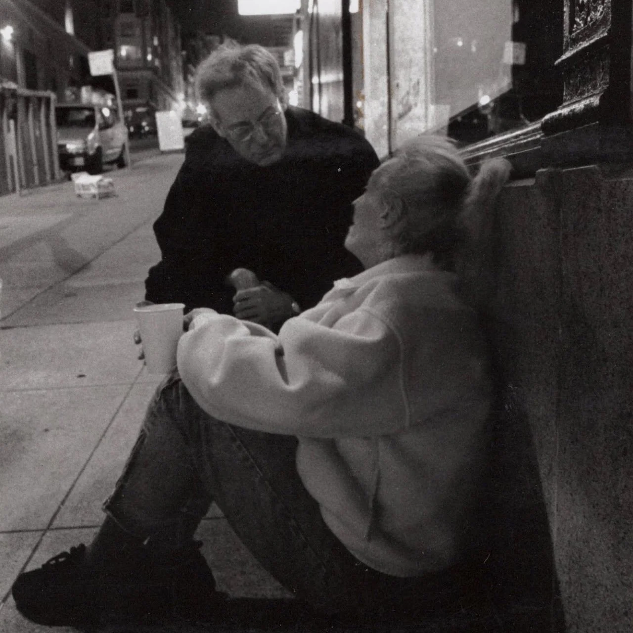 A young person sitting on the sidewalk at night, talking to an elderly woman with gray hair who is resting against a wall. The young person is holding a cup and a microphone, and the elderly woman appears to be listening attentively.