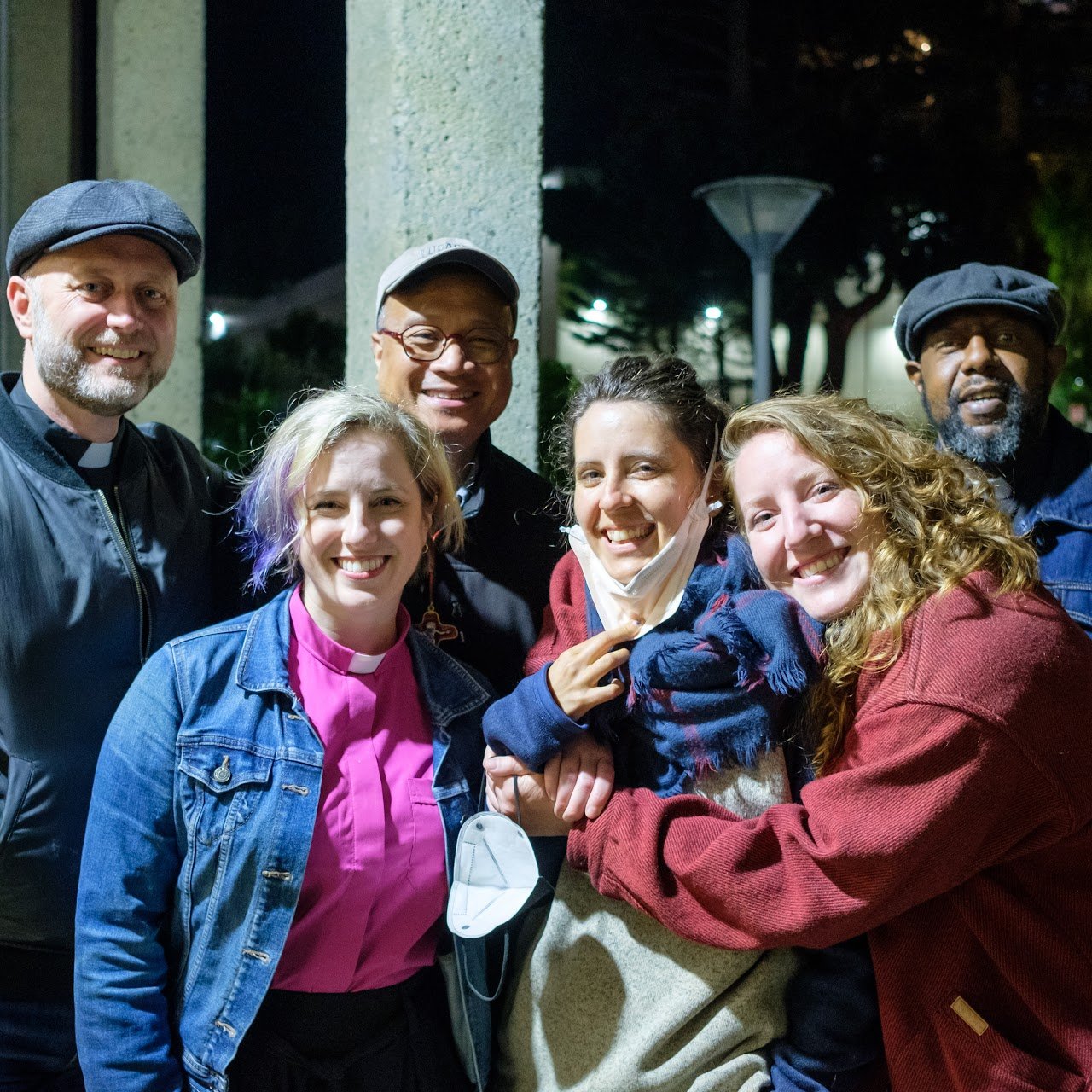 Group of six people smiling and hugging outside at night, some wearing jackets and face masks.