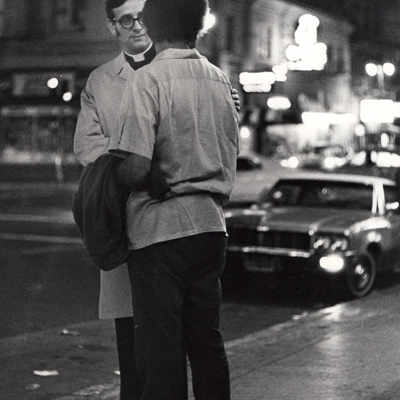 A black and white photo of a person dressed as a priest talking to a woman on a city street at night, with cars and neon signs in the background.
