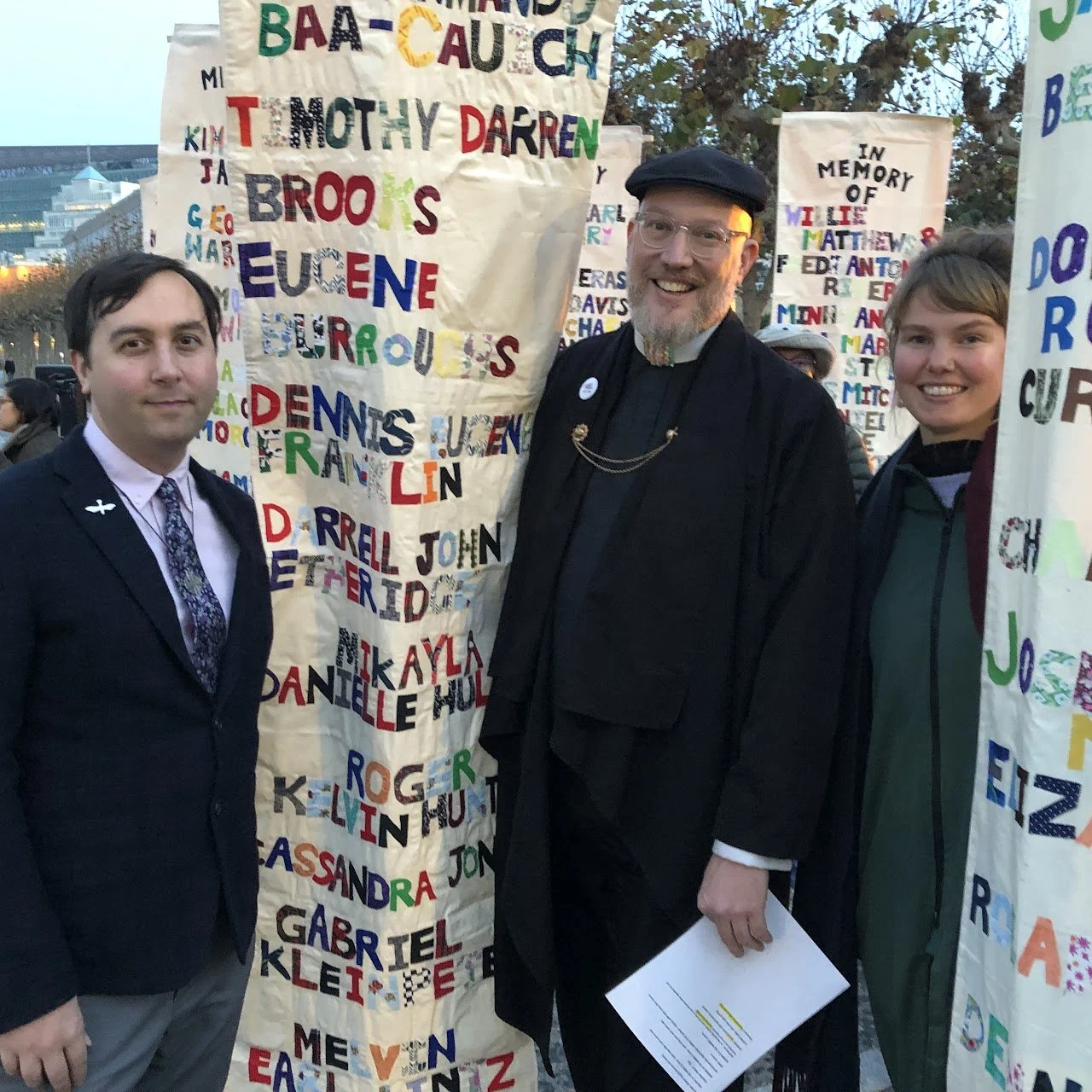 Three people standing outdoors among colorful handmade posters with names and messages, smiling at the camera.
