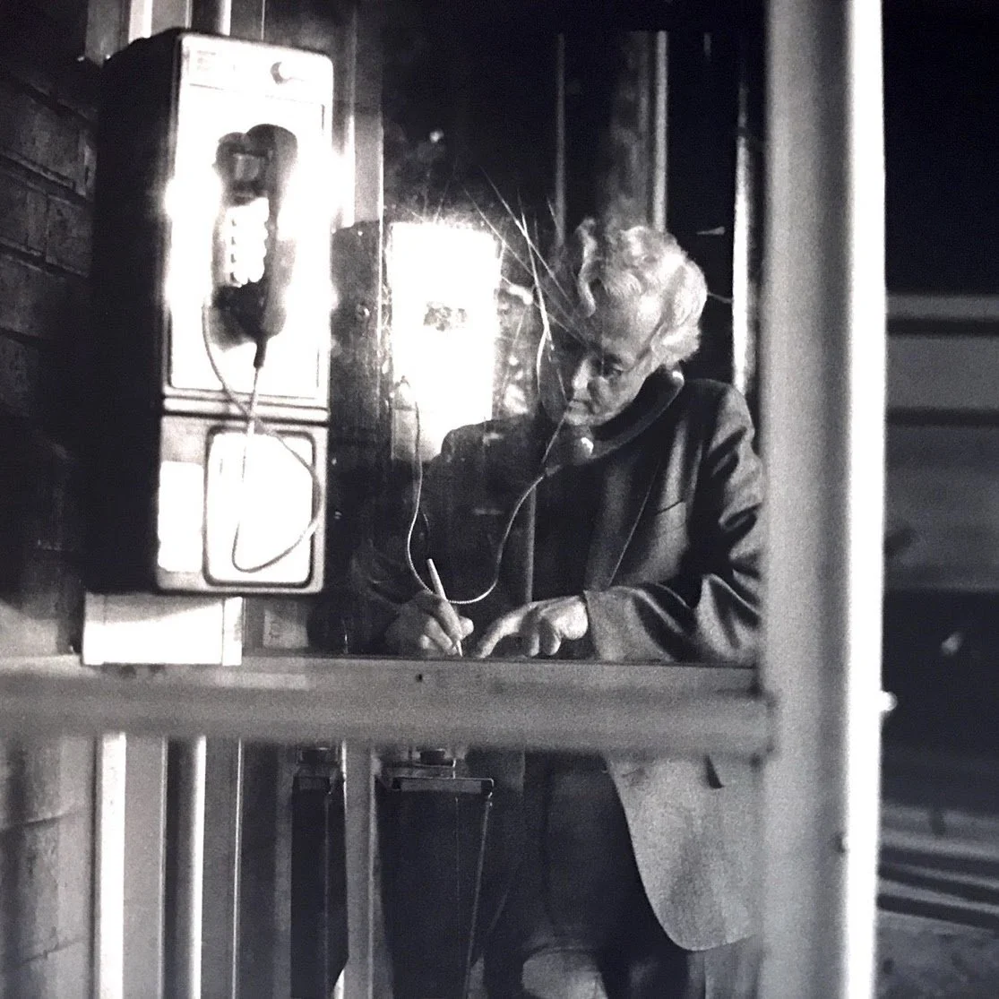 Black and white photo of an elderly woman wearing glasses, writing with a pen on paper at a table, with an old telephone on the wall in front of her.