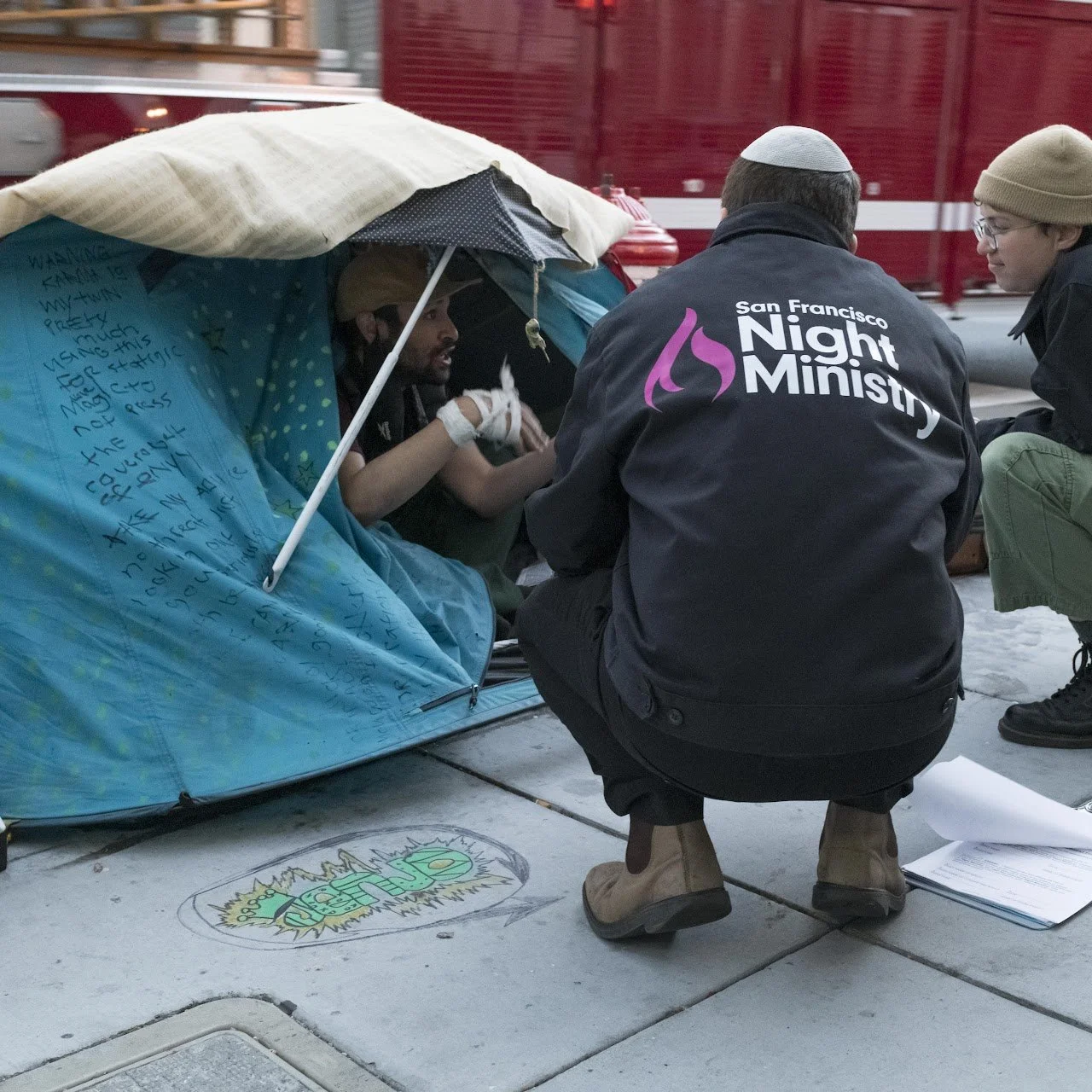 A man inside a blue tent on the sidewalk, talking with two people in dark jackets. One jacket has 'San Francisco Night Ministry' printed on the back. The tent has handwritten notes on its inside wall.
