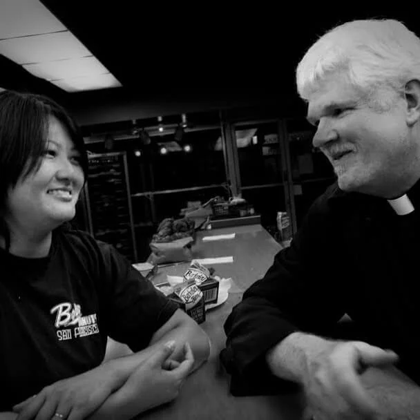 A woman and a priest smiling and shaking hands at a bar or counter in a restaurant or cafe, with drinks and snacks on the counter behind them, in a black and white photo.