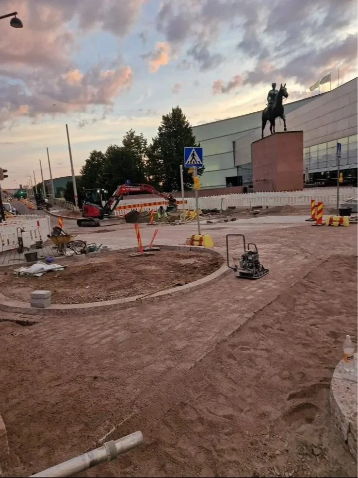 Kuvassa rakennustyömaalla on kaivinkone ja työskenteleviä ihmisiä. Taustalla on julkinen taideteos eli ratsastajahahmoa esittävä patsas, ja taivaalla on pilviä auringonlaskussa.