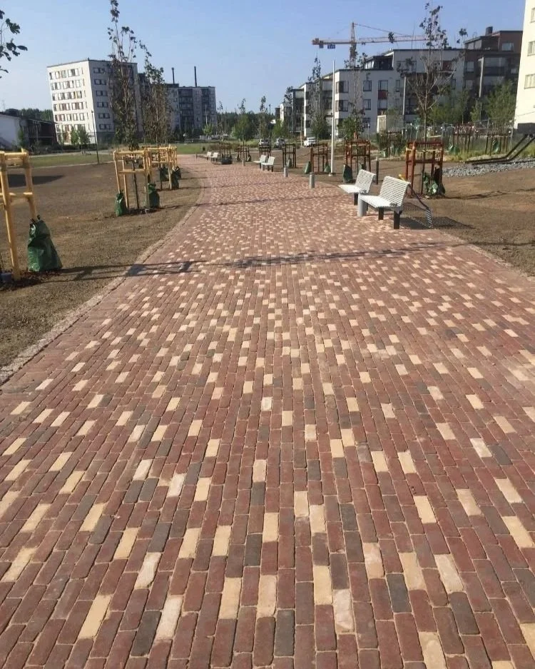 Kuvassa on uusi kävelytie, jonka päällysteenä on erilaisia punaisia ja beigejä tiiliä. Taustalla on keskieurooppalaisia kerrostaloja ja rakennustelineitä. Kävelytie on osittain vielä keskeneräinen ja ympäristöä valmistellaan.