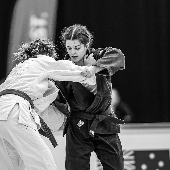 Two young women practicing judo on a mat, wearing traditional judo uniforms. One is in a white gi with a black belt, and the other is in a black gi. They are engaged in a close fighting stance, focusing on each other in an indoor dojo.