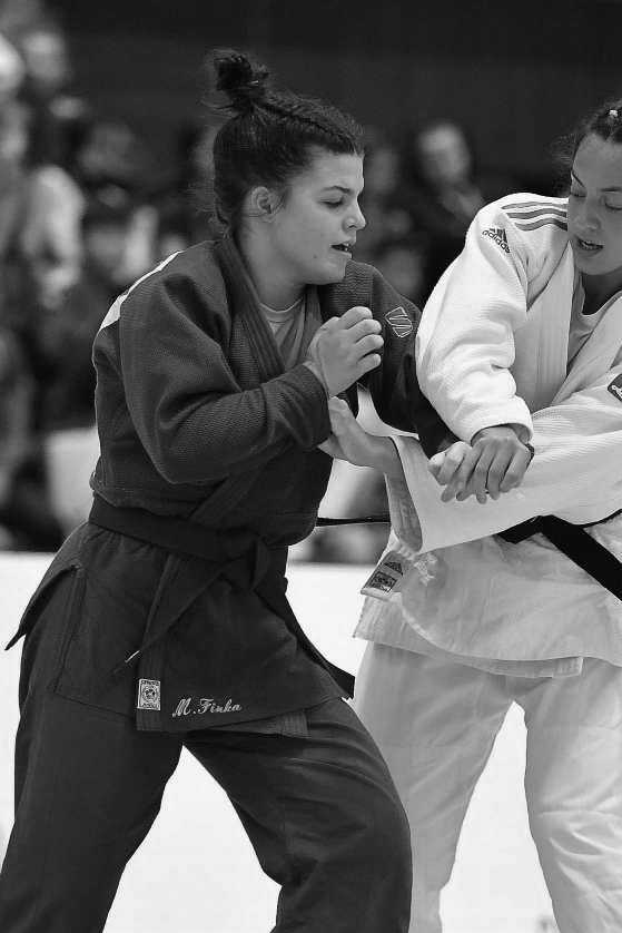 Two female judo practitioners during a match, with one gripping the other's judo gi while focusing on her technique, in a competitive setting.