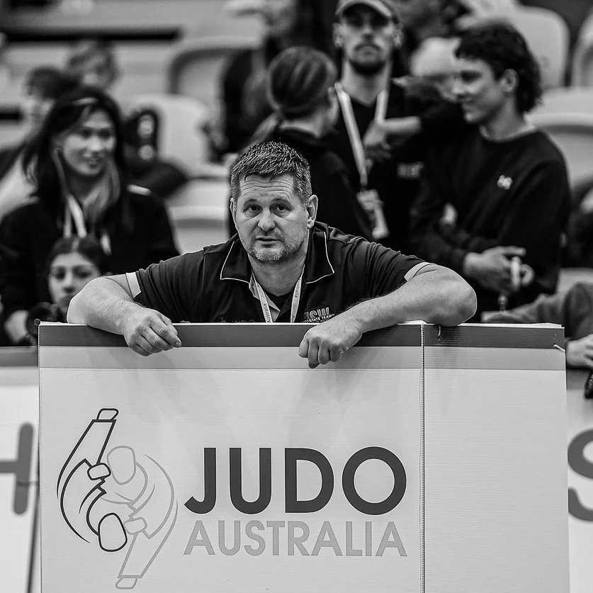 A man leaning on a sign that says "Judo Australia", with several people in the background at a judo event or competition.