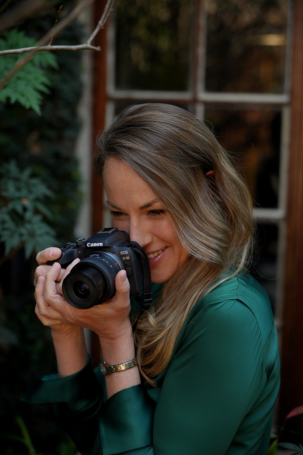 A woman with long, wavy blonde hair wearing a green shirt, smiling as she looks through the viewfinder of a Canon EOS R10 camera, in an outdoor garden setting.