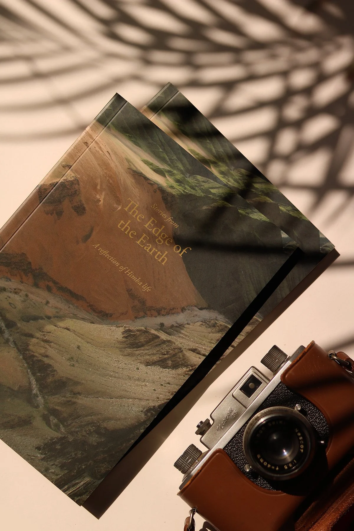 A book titled 'Stories from The Edge of the Earth' resting on a white surface. Shadows of a lattice pattern are cast over the book. Next to the book, there is a vintage camera with a leather strap and a small metallic object.