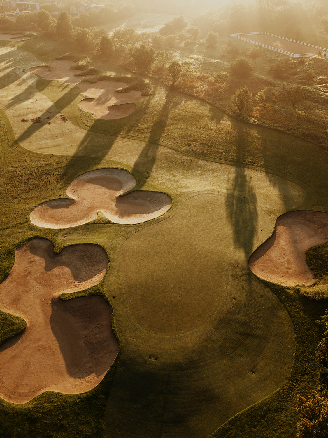 An aerial view of a golf course at sunset, showing sand bunkers, green fairways, and trees casting long shadows.