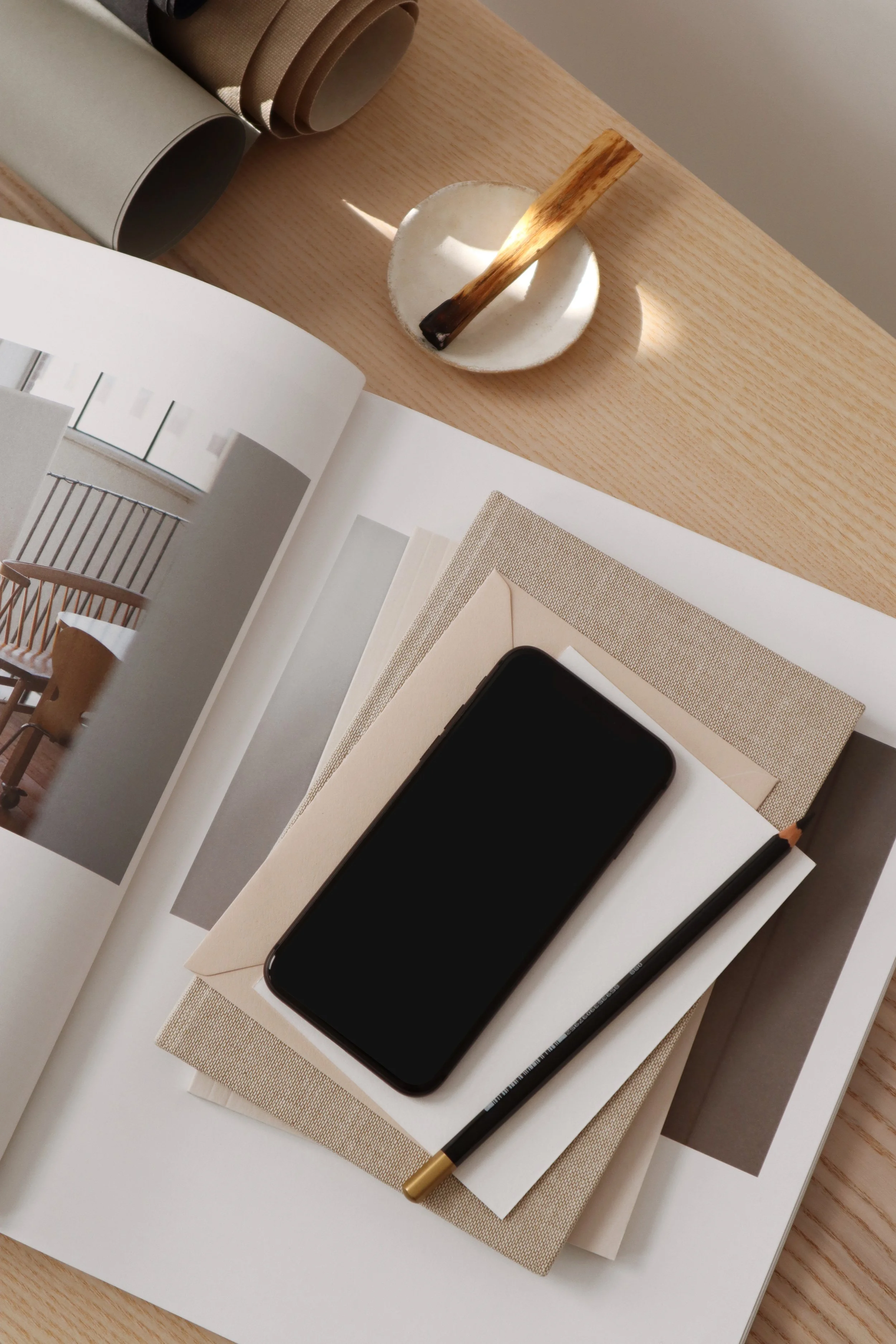 Open magazine showing furniture design images, placed on a beige textured folder, with a black smartphone, a black pencil, a beige textured folder, a white basket with a wooden stick, and two rolls of wrapping paper on a light wood table.