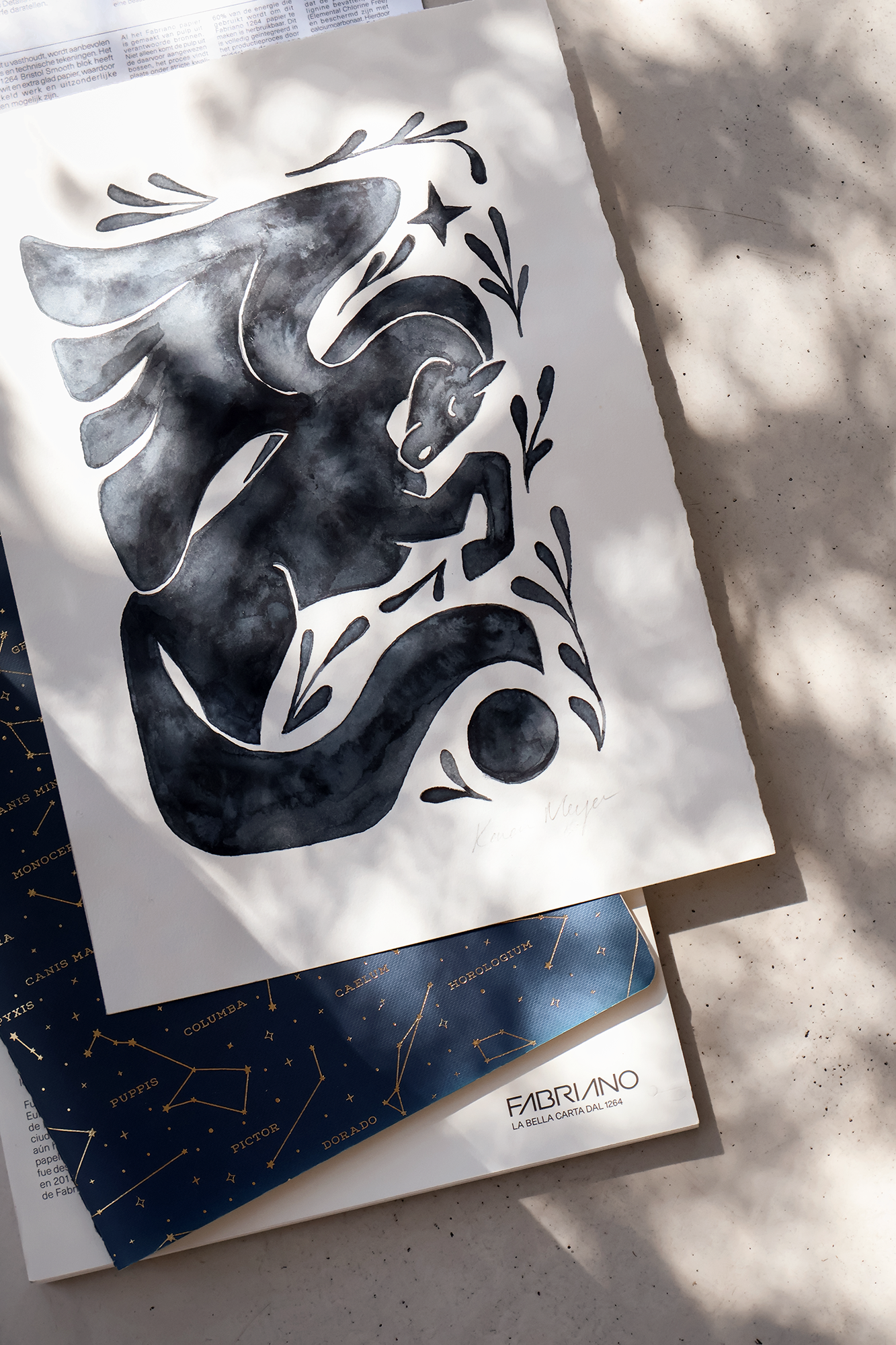 Black and white artistic drawing of a horse with decorative foliage around it, signed by Kent Meyer. The drawing is on a piece of paper placed on top of dark blue paper with gold constellation designs and labels of constellations, with light shadows and some fallen leaves on the surface.