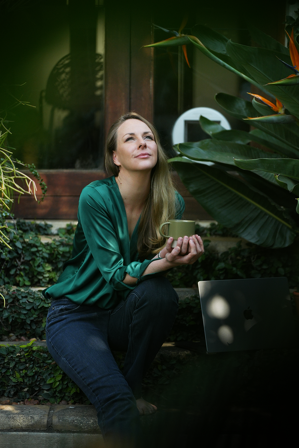 A woman with long blonde hair sitting outdoors on stone steps, holding a coffee mug and looking thoughtfully upward amidst lush greenery and large tropical leaves.