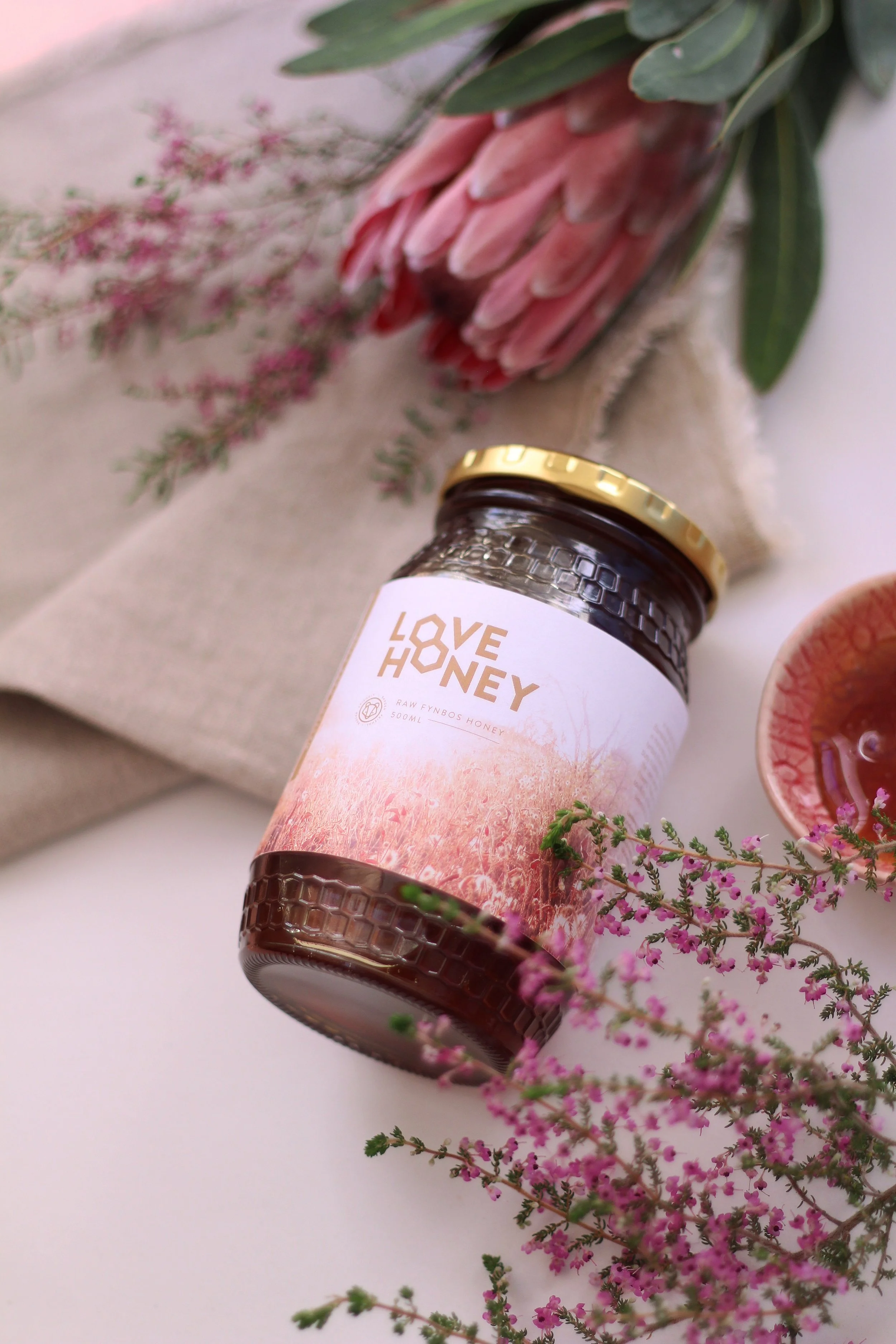 A jar of Love Honey with pink floral artwork on the label, surrounded by pink flowers and green leaves, on a white surface.