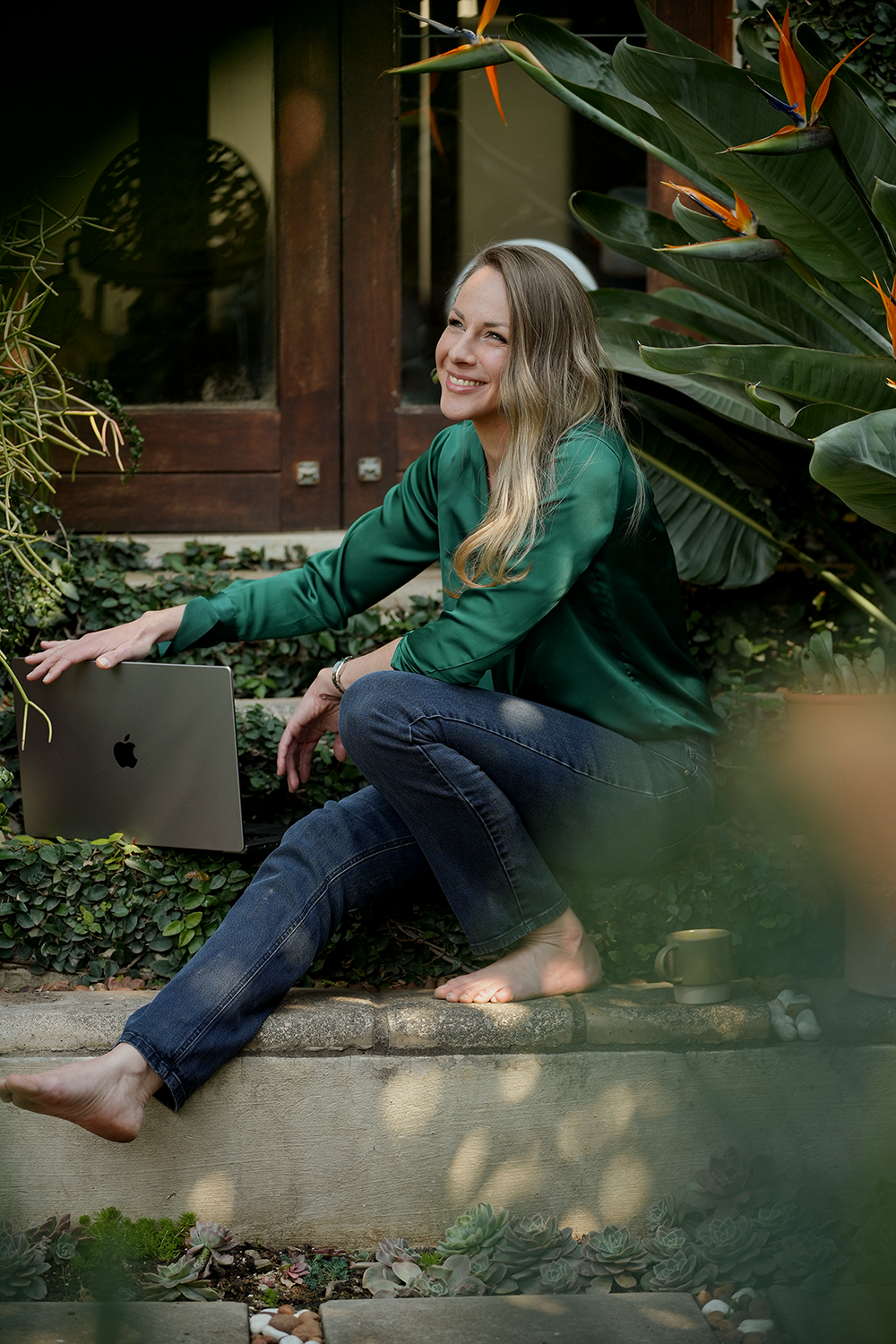 A woman sitting outdoors on a garden ledge, smiling and reaching toward a closed laptop, surrounded by lush green plants and succulents, with a mug beside her.