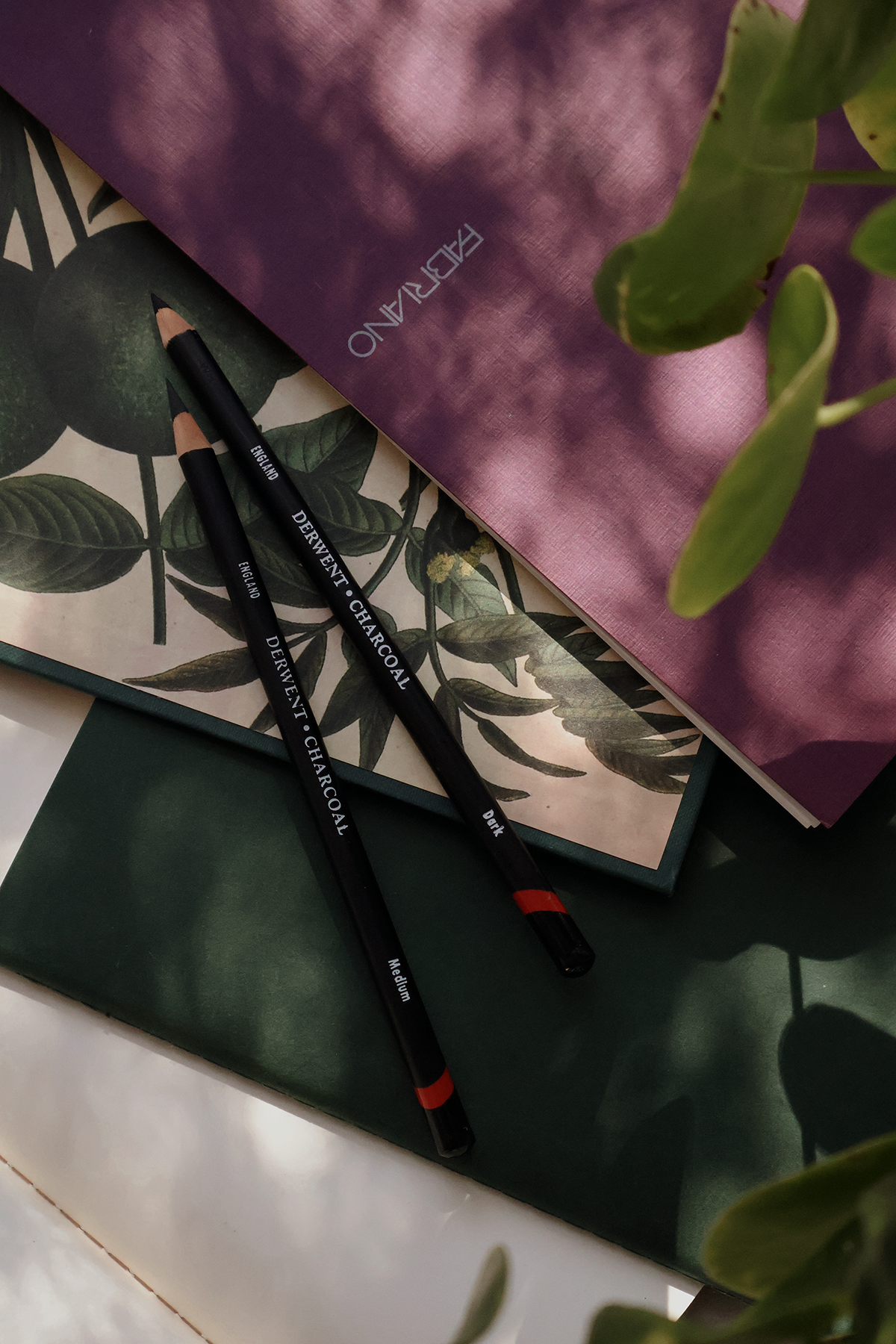 A close-up of two black pencils with white text, placed on botanical printed paper. A purple and green textured book or magazine cover is partially visible, with green leaves and a sprig of a plant nearby.