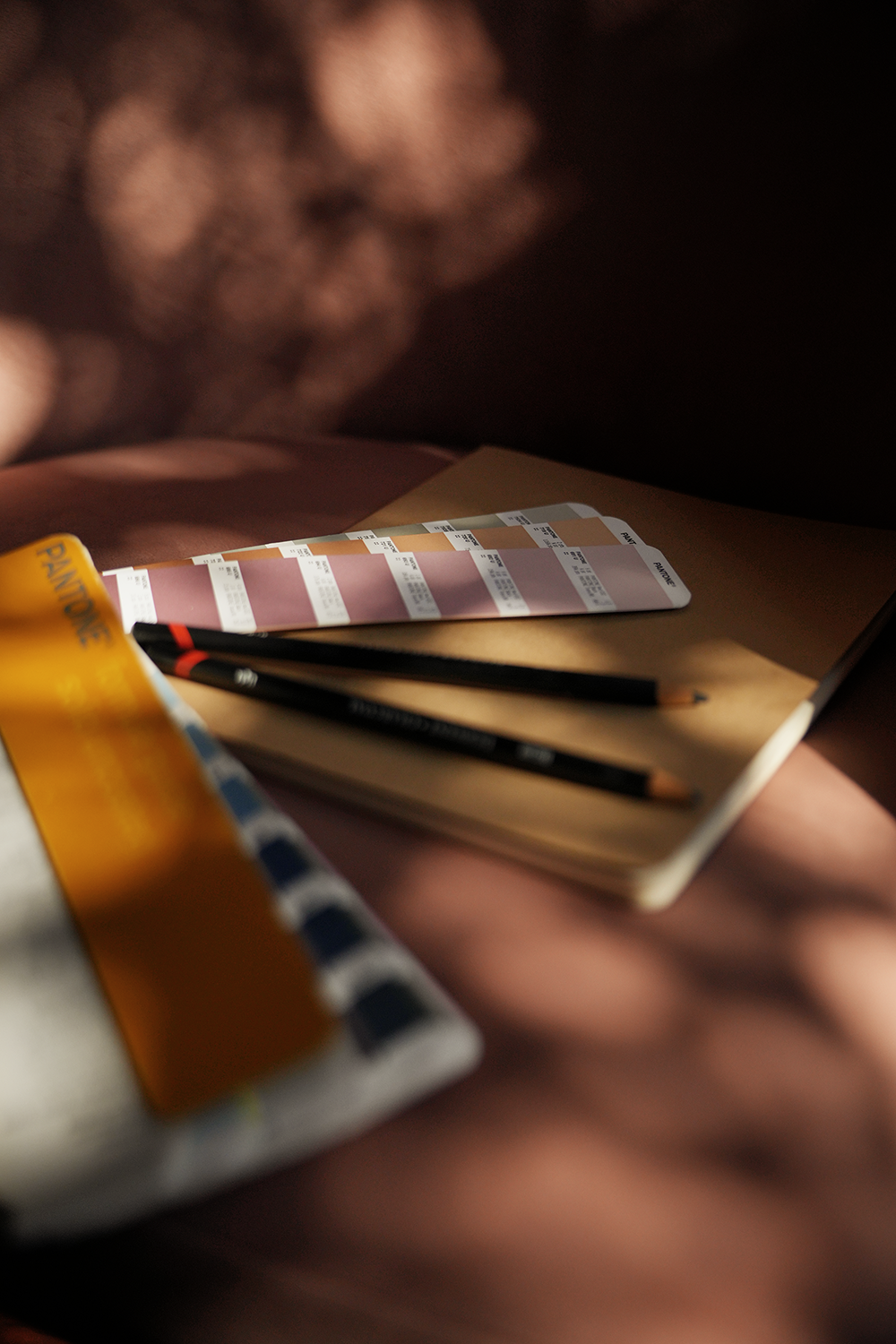 A sketchbook, two colored pencils, and a color swatch chart on a wooden surface with shadows.
