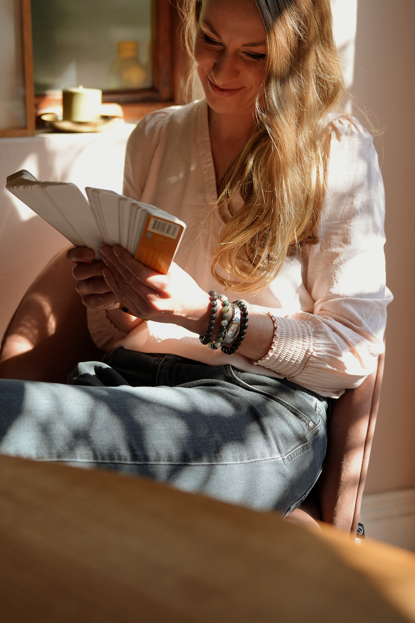A woman with long blonde hair smiling and looking down at a fan of postcards or photographs she is holding in her hands, seated in a room with sunlight.