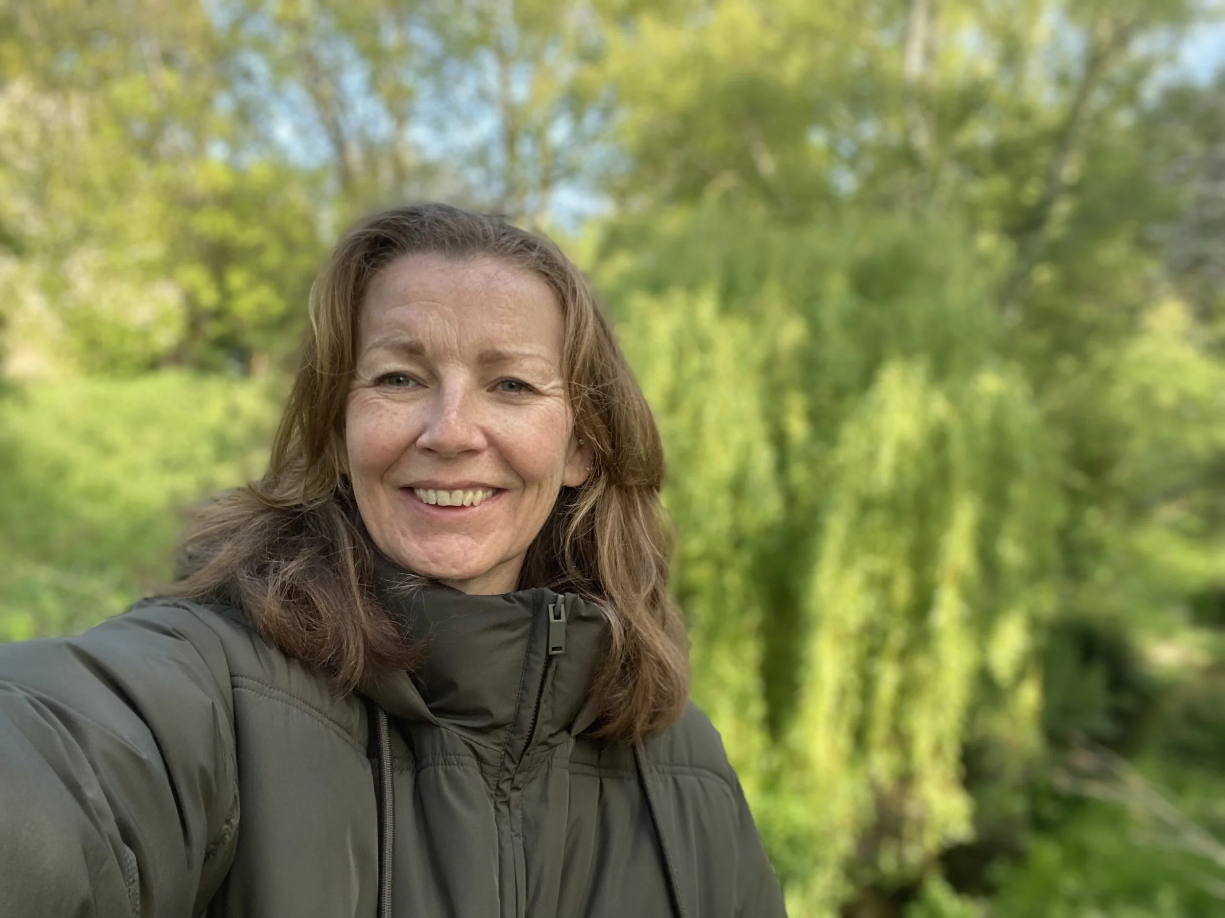 Louise standing outdoors in a green, leafy setting, wearing a padded jacket and smiling softly at the camera.