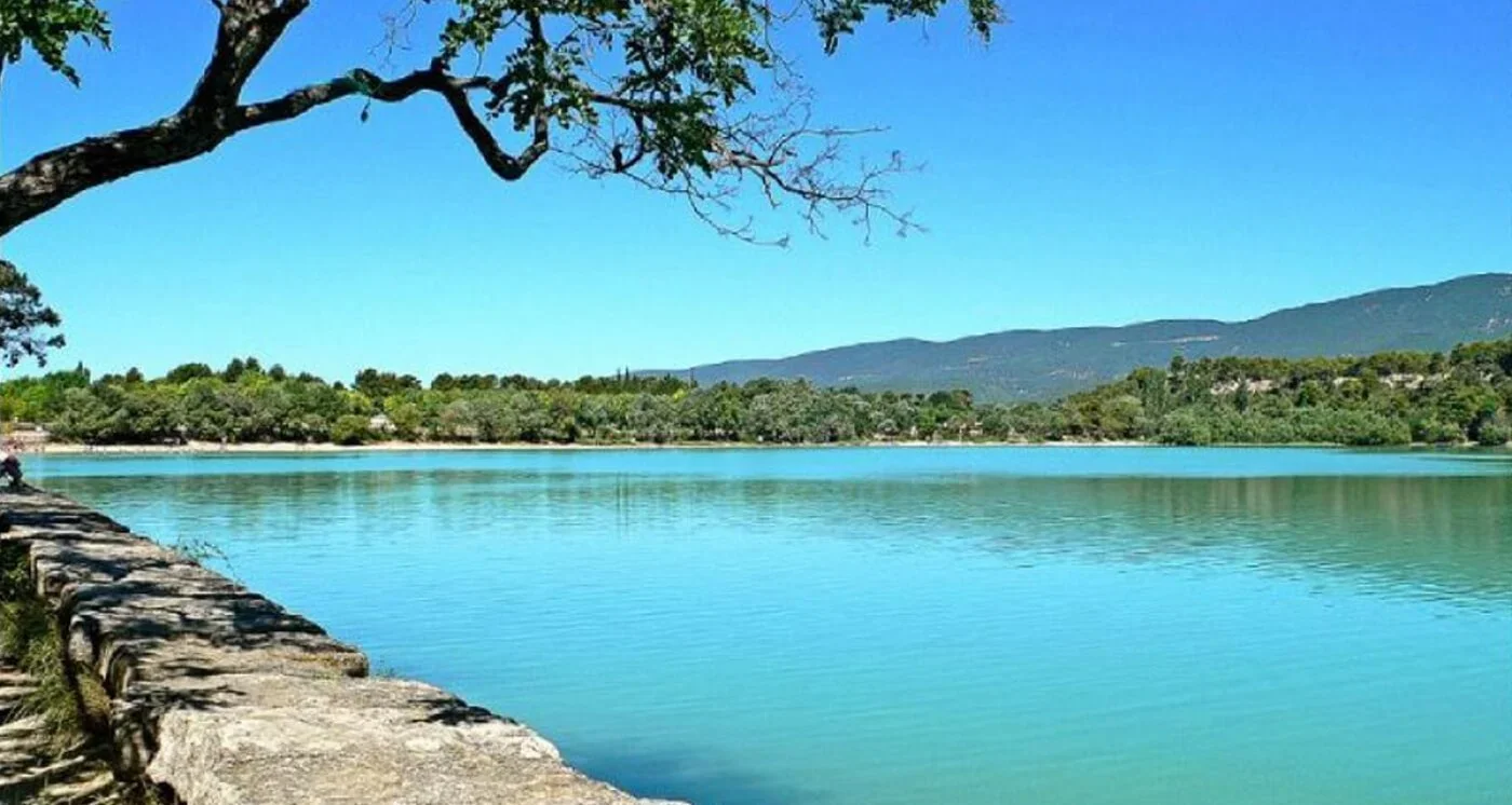 A peaceful lake scene with clear blue water, a stone wall along the shore, overhanging tree branches, green trees on the opposite bank, and distant mountains under a bright blue sky.
