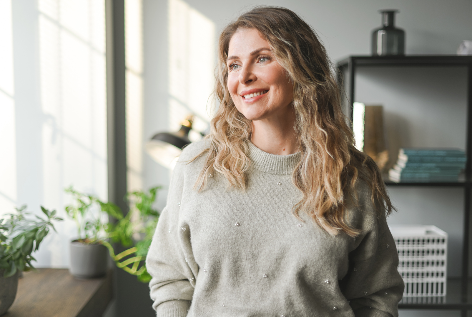 A woman with light brown curly hair smiling in an indoor space with sunlight and houseplants.