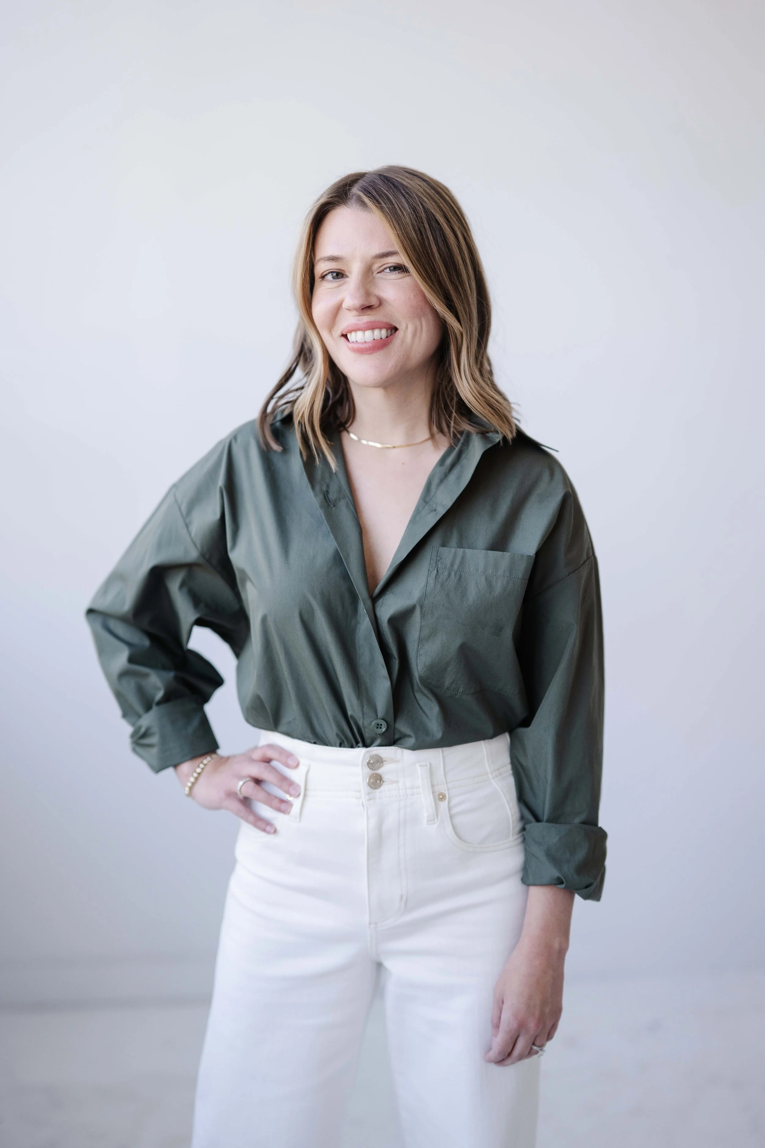 A woman with shoulder-length brown hair wearing a dark green button-up shirt and high-waisted white jeans, standing against a plain white background, smiling with one hand on her hip.