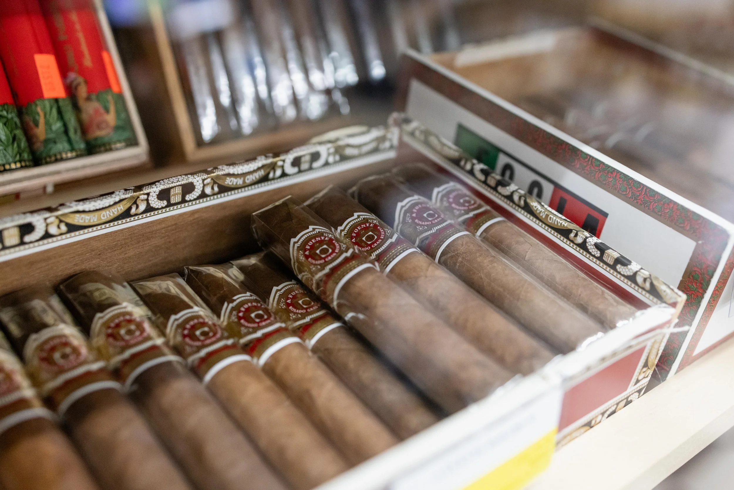 Close-up of a box of cigars with a partially opened lid, showing cigars wrapped in plastic with red and white labels inside a decorative box.