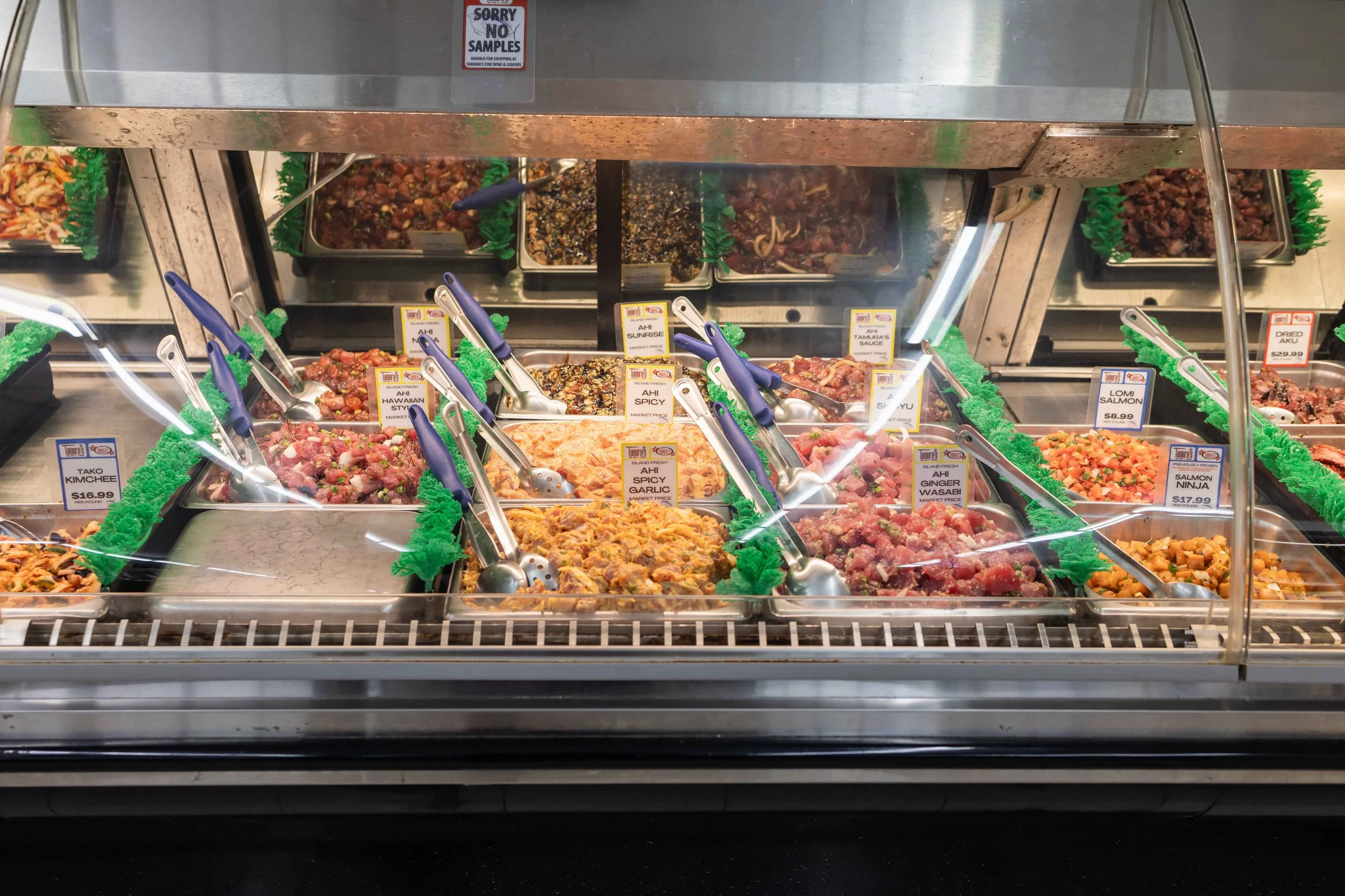 Display of various prepared Asian-style meat dishes in a glass showcase at a grocery store, with labeled prices and serving tongs.